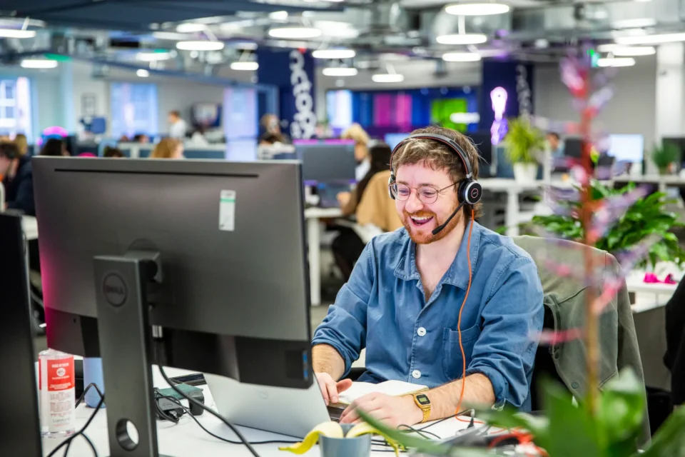 A smiling man wearing a headset works at a computer in a busy office.